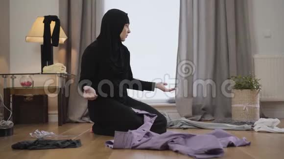 绝望的穆斯林妇女坐在地板上环顾四周散落的衣服的肖像东部家庭主妇视频的预览图
