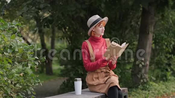 戴着帽子穿着毛衣的年轻红发女郎在长椅上看书喝着热咖啡视频的预览图