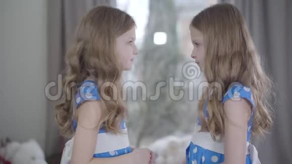 侧视中镜头愤怒的小白种人女孩骂她的双胞胎妹妹穿着蓝色点缀礼服的漂亮兄弟姐妹视频的预览图