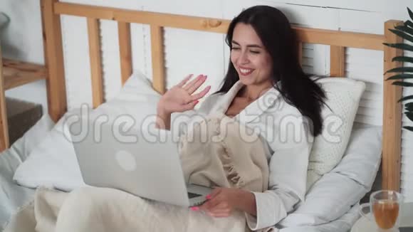 穿着浴袍的年轻女子的肖像躺在床上并在笔记本电脑上进行视频聊天视频的预览图
