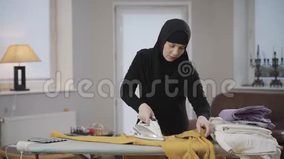 穆斯林妇女集中在头巾熨烫黄色套头衫的肖像家庭主妇在室内做家务父权制视频的预览图