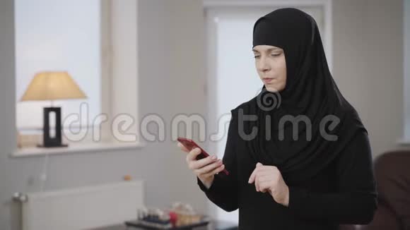 年轻穆斯林妇女在智能手机上使用社交媒体的侧视微笑的现代女士戴着头巾在家休息视频的预览图