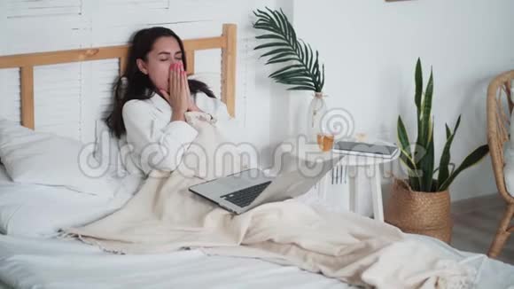 疲惫的女人坐在床上在家里用笔记本电脑工作伸懒腰打哈欠视频的预览图