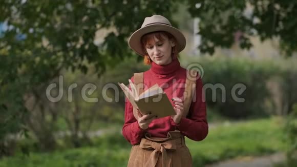 年轻迷人的红头发女人戴着帽子和毛衣在一个自然公园看书秋天凉爽的一天视频的预览图