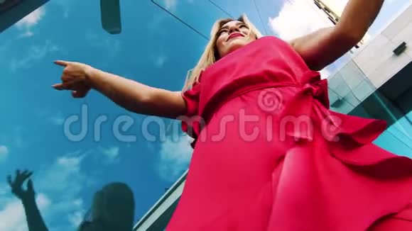 金发女人穿着红色连衣裙靠玻璃墙自由舞蹈的低角度视频的预览图