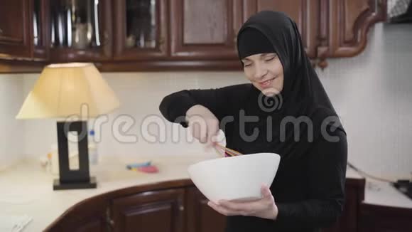 中镜头的美丽穆斯林妇女穿着传统头巾看着相机和闪光灯现代东方女人做饭视频的预览图