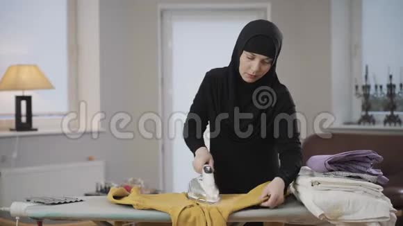 勤劳的穆斯林家庭主妇穿着黑色头巾在家熨黄色毛衣年轻漂亮的女人做家务东部视频的预览图