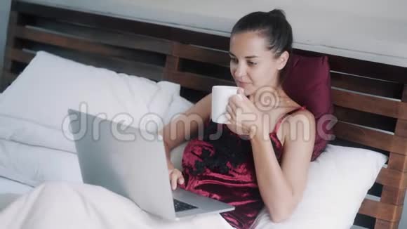 女人在笔记本电脑上工作在舒适的床上喝咖啡视频的预览图