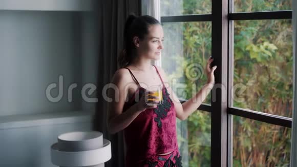 穿着雅致睡衣的快乐女人在窗边喝果汁视频的预览图