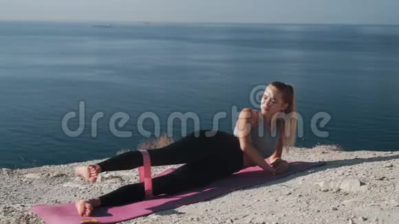 女孩躺在垫子上用橡皮筋做运动训练她的臀部和身体视频的预览图