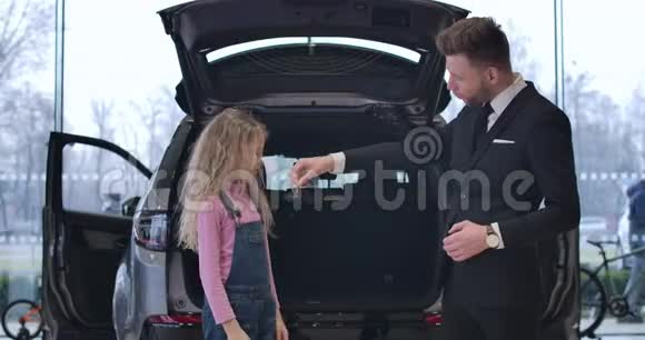 年轻的白种人给一个留着卷发的金发小女孩车钥匙汽车经销商和可爱的客户站在视频的预览图