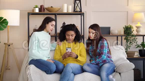 悲伤的多种族女孩在家中浏览智能手机上的社交媒体视频的预览图