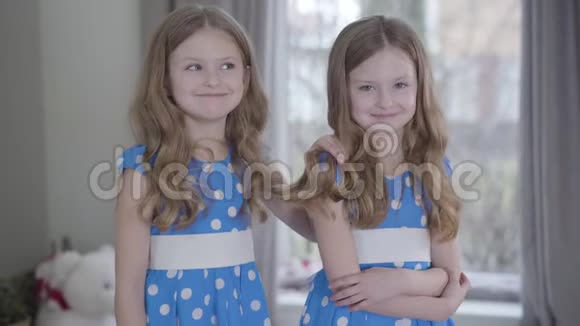 笑容满面的黑发白种人双胞胎姐妹穿着类似点缀的蓝色连衣裙在家里摆姿势积极的小妹妹视频的预览图