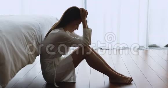 穿着睡衣坐在靠近床的地板上的年轻沮丧的女人视频的预览图