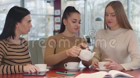 年轻的黑发白种人女人把茶倒在杯子里和女性朋友交谈三个美丽的女孩休息视频的预览图