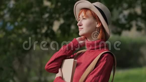 年轻迷人的红头发女人戴着帽子和毛衣在自然公园的长凳上看书秋天凉爽的一天视频的预览图