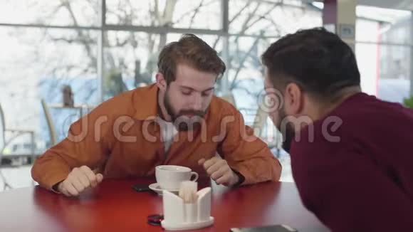 积极的年轻白种人的肖像穿着芥末衬衫在咖啡馆里和胡子朋友交谈两个男性朋友视频的预览图