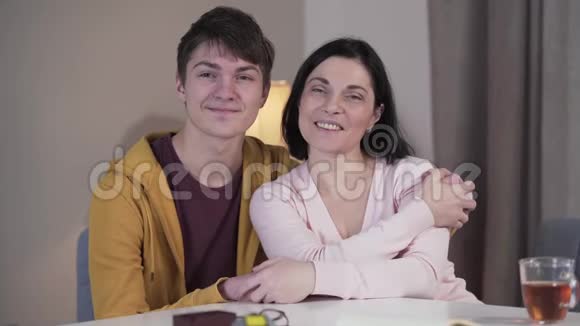 中镜头的快乐白种人的儿子和母亲看着相机微笑着坐在室内的桌子少年儿童视频的预览图