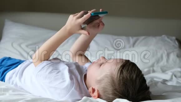 可爱的小男孩蹒跚学步玩手机游戏躺在床上拿着智能手机视频的预览图