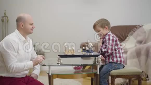 侧面看成年白种人男子教小儿子下棋小可爱的男孩在家和爸爸一起学习团结视频的预览图