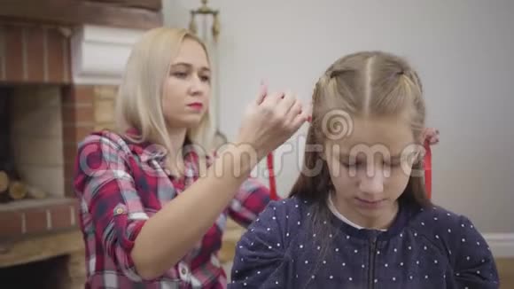 摄像机放大了金发白种人成年女性为大女儿做发型十几岁的女孩编织妹妹视频的预览图