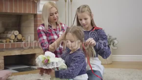镜头接近美丽的白种人女孩玩娃娃作为她的姐姐和母亲编织辫子快乐视频的预览图