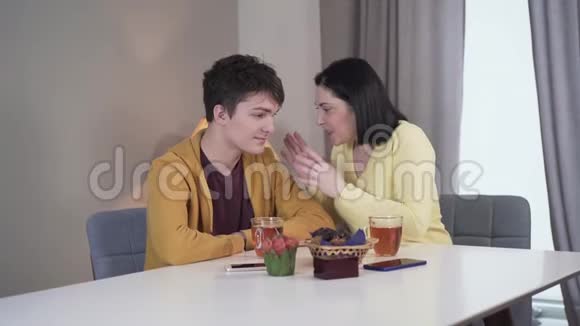 中镜头快乐的年轻白种人女人讲笑话的十几岁的男孩坐在桌子上室内Brunette视频的预览图