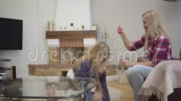 侧面观看欢快的白种人女孩在家中捕捉肥皂泡微笑的金发女人吹泡泡给女儿视频的预览图