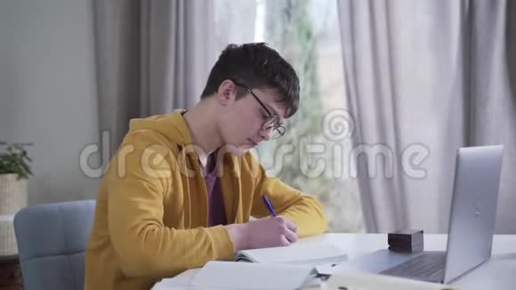相机从左到右移动在年轻的白种人男孩在眼镜写作和使用笔记本电脑智能书呆子视频的预览图