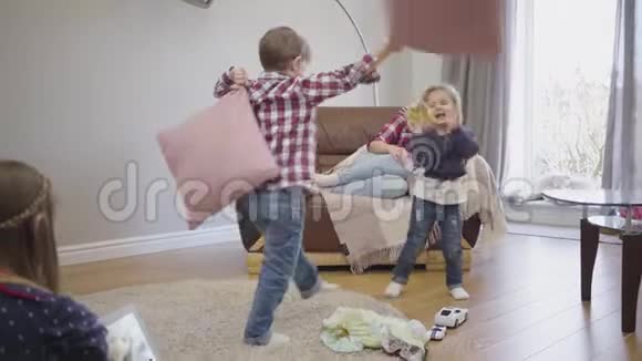 有趣的小白种人的孩子们抱着枕头到处乱跑他们的母亲坐在沙发上和长辈在一起视频的预览图