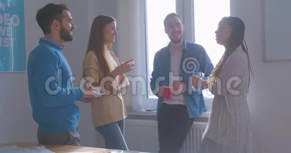 四个不同种族的同事在办公室喝咖啡休息谈话讨论阳光房休息笑话视频的预览图