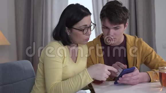 白种人成年女性在十几岁的儿子帮助下学习使用电话的肖像自信的小男孩视频的预览图