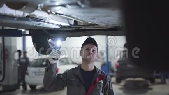 严肃的白人男子穿着工作服戴着帽子用手电筒在车底下行走男性成人汽车修理师视频的预览图