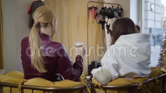 女性朋友坐在时尚精品店里带着咖啡带着微笑的金发女人走出更衣室视频的预览图