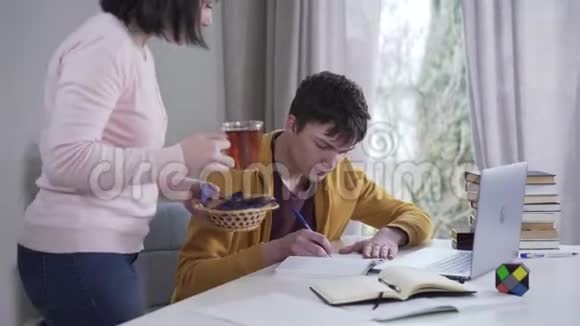 细心的白人母亲给在餐桌上努力学习的十几岁男孩端上茶和糖照顾黑发女人视频的预览图