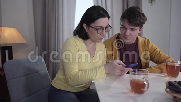 年轻的黑发白种人男孩教成年母亲戴眼镜使用智能手机有病人的儿子帮女人交易视频的预览图