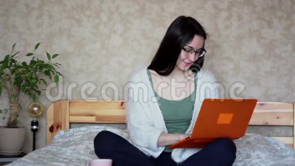 年轻漂亮的女孩正在用她的笔记本电脑工作坐在家里的床上年轻可爱的女孩正坐在床上打字视频的预览图