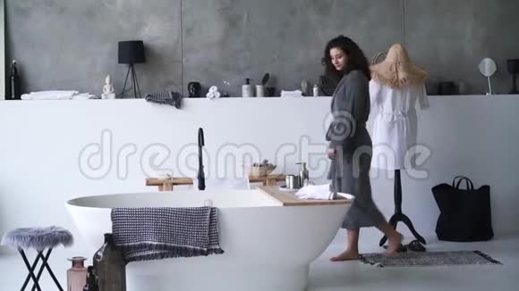 年轻的黑发女人穿着浴袍在浴缸里检查水温漂亮的白种人女孩在大浴室里散步视频的预览图