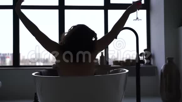 年轻女人在浴缸里喝酒的剪影开朗的白人女孩在大窗户前的浴室里休息视频的预览图