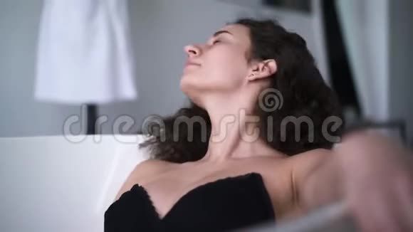 躺在浴缸里的棕色眼睛黑发女人的肖像一位美丽的白人女孩卷发洗澡视频的预览图