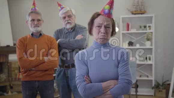 一幅高加索成年女性沮丧的肖像双手交叉地站在养老院其他居民戴着派对帽视频的预览图