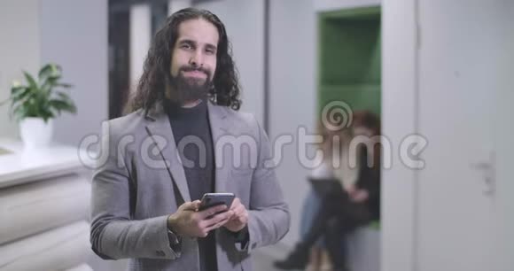 英俊的中东男人在智能手机键盘上打字看着相机微笑着的肖像自信办公室视频的预览图