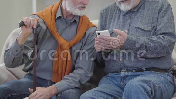 两个无法辨认的白人老人使用智能手机和谈话快乐的男性退休人员在护理沙发上聊天视频的预览图