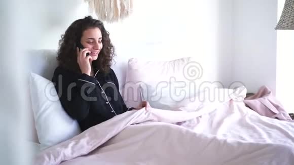 年轻的白种人女人在卧室里打电话家中躺在床上的快乐卷发黑发女孩的肖像视频的预览图