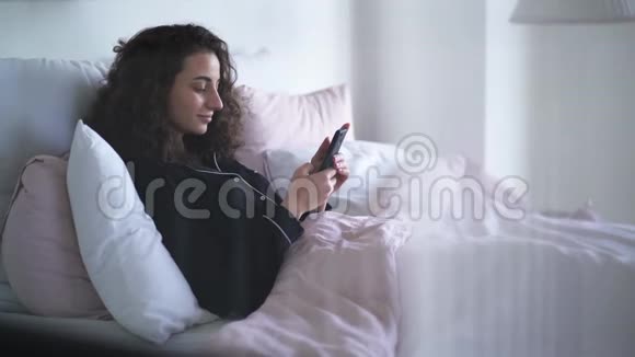 年轻积极的女人用智能手机笑躺在床上的黑发卷发白种人女孩的侧景视频的预览图