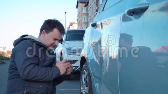 男子正坐在地上的停车场上的街道上的电话为保险公司在车门上拍摄凹痕的照片他是视频的预览图