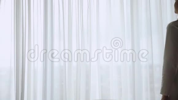 年轻的黑发女人穿着睡衣打开长长的浅色窗帘视频的预览图