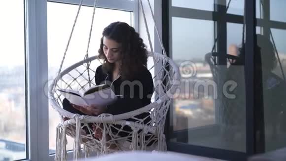 年轻的高加索女人坐在挂着的扶手椅上看书热衷于文学的聪明女孩的肖像视频的预览图