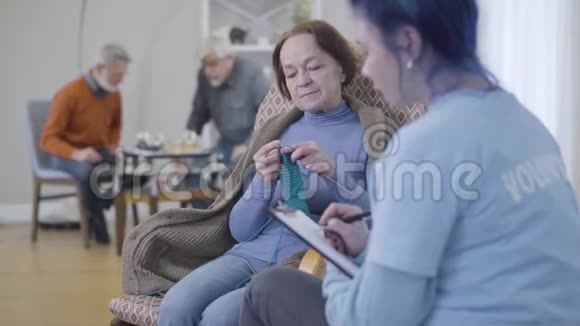 白种人模糊的志愿者在养老院和女性退休人员交谈一位老年妇女在编织和示意不要视频的预览图