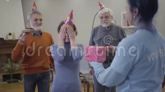 兴奋的老白种人女人打开眼睛作为志愿者和老年男子带来礼物和生日蛋糕的肖像视频的预览图